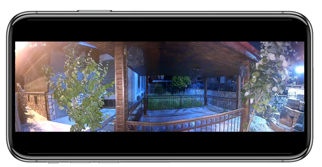 Bir verandadan çekilen gece görüntüsü, akıllı telefon ekranından görüldüğü gibi, kapalı bir veranda alanını, metal korkuluğu, yeşilliği ve aydınlatılmış bir bahçeyi gösteriyor.
